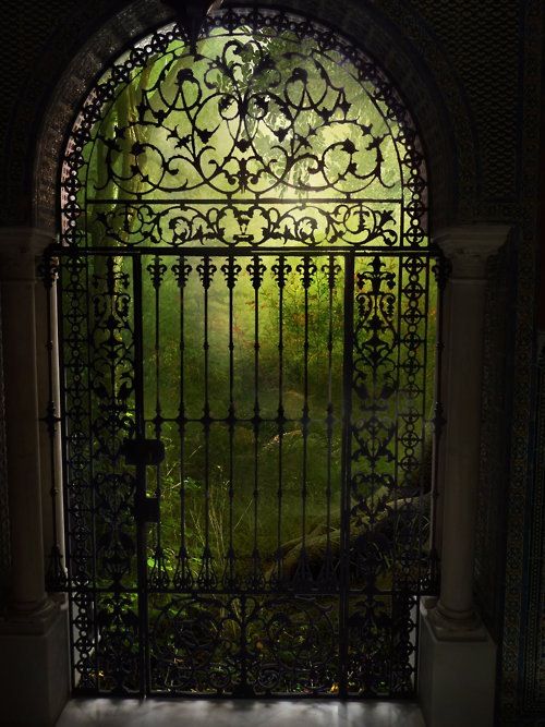 an iron gate is open to reveal a lush green park in the distance, with trees and bushes on either side