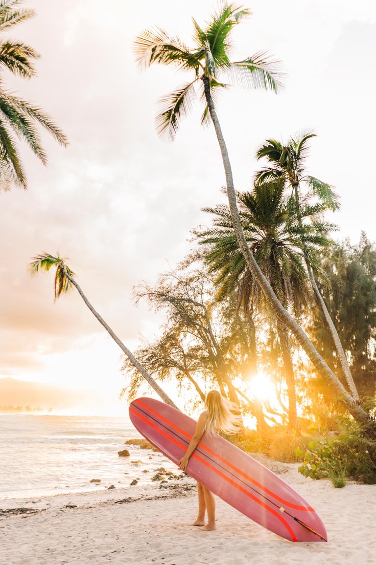 a woman holding a red surfboard on top of a sandy beach next to palm trees