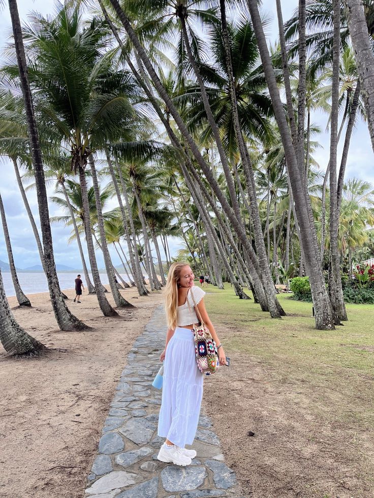 a woman walking down a stone path between palm trees and the ocean on a sunny day