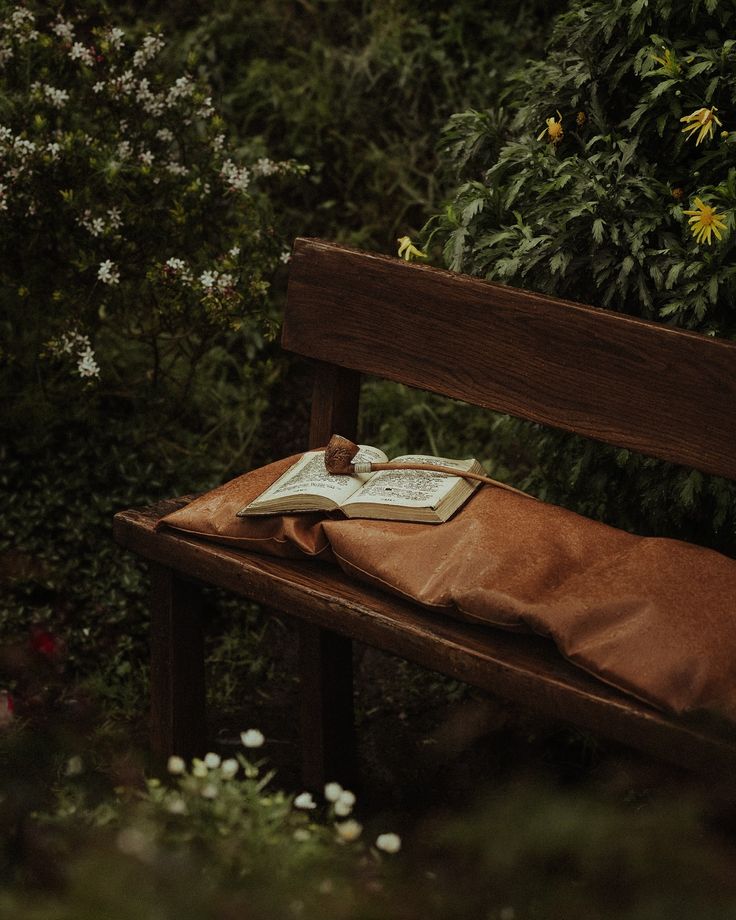 an open book sitting on top of a wooden bench in front of some bushes and flowers