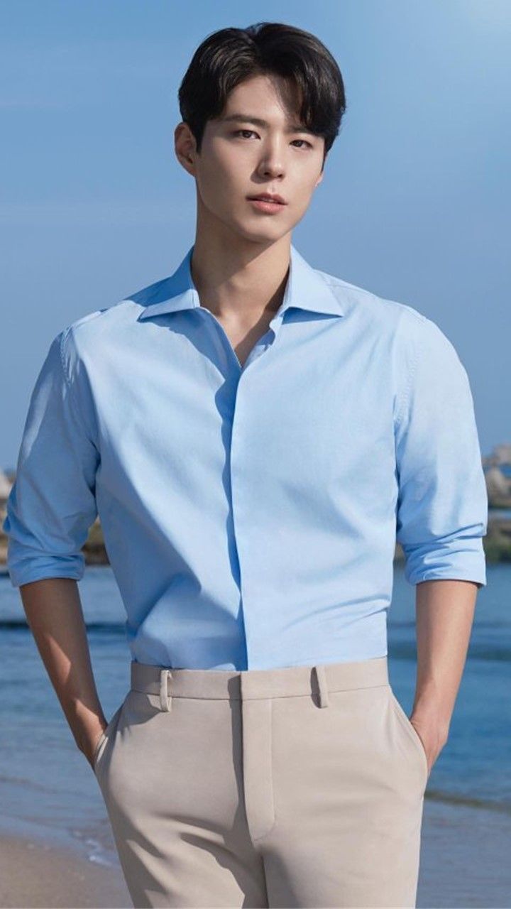 a young man standing on top of a beach next to the ocean wearing a blue shirt and khaki pants