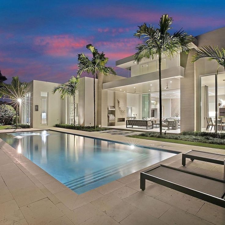 an outdoor swimming pool with lounge chairs and palm trees in front of the house at dusk