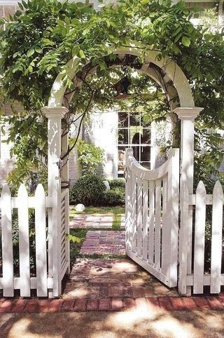 a white picket fence with an arch over it and trees growing on the top, in front of a house