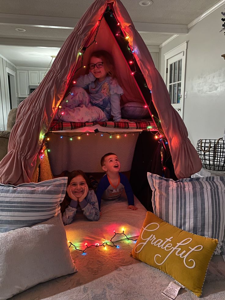 two children are laying on the floor in a tent with christmas lights and pillows around them