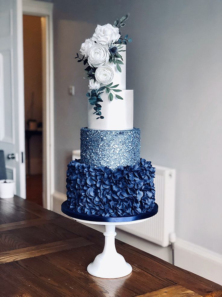 a three tiered blue and white wedding cake with flowers on the top, sitting on a wooden table