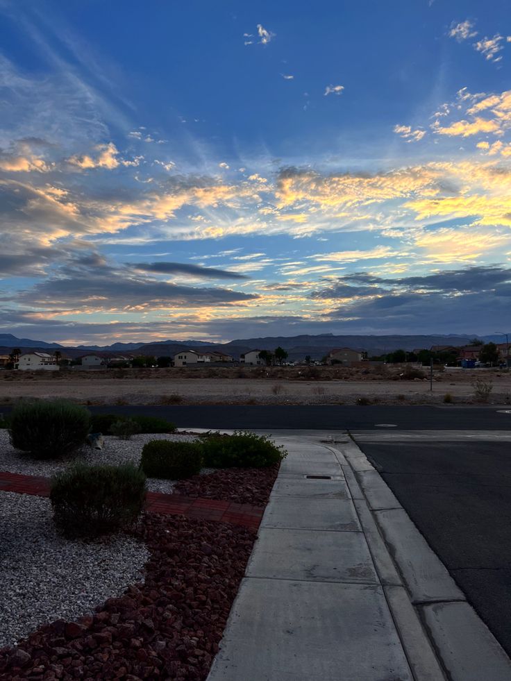 the sun is setting over an empty street in front of some houses and mountains with clouds