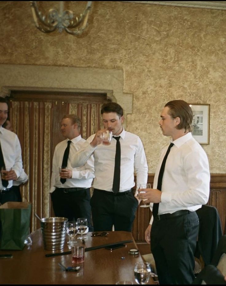 four men in formal wear standing around a table drinking wine and talking to each other