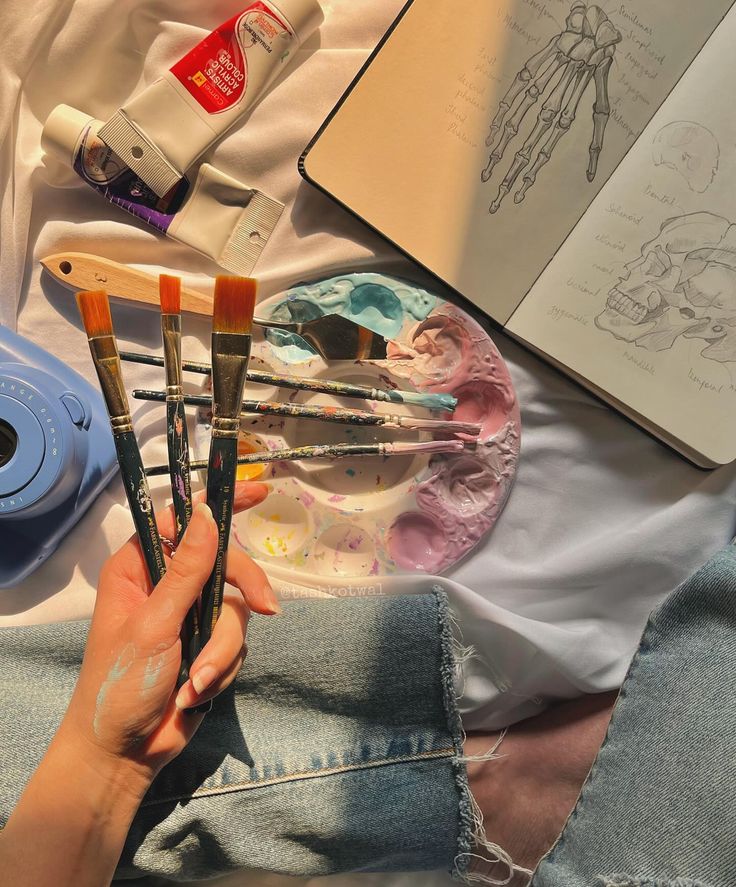 someone is holding paintbrushes in front of some art supplies on a bed with an open book