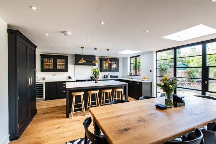 a kitchen with black cabinets and wooden table in the foreground is an open floor plan