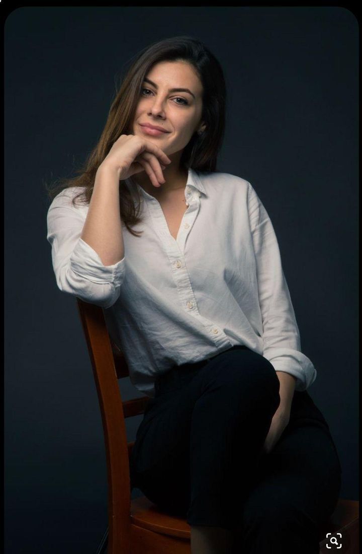 a woman sitting on top of a wooden chair in front of a dark background with her hand under her chin