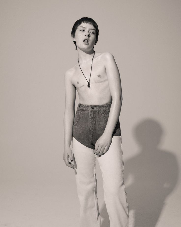 black and white photograph of young boy with no shirt standing in front of grey background