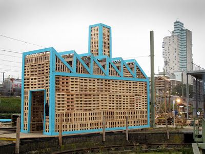 a building made out of wooden pallets sitting on top of a train track next to tall buildings