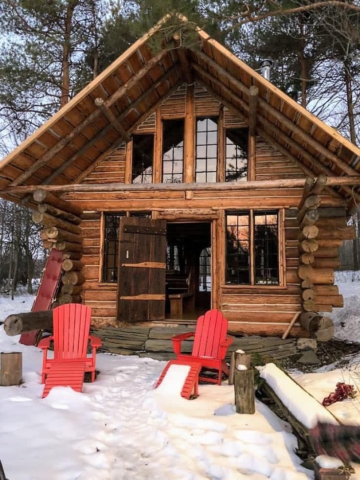 two red chairs sitting in front of a log cabin with snow on the ground and trees