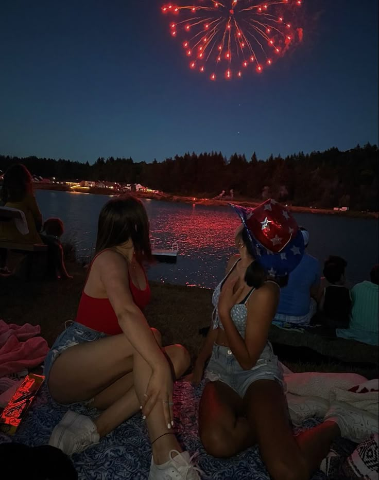 two women sitting on the ground next to each other watching fireworks in the night sky