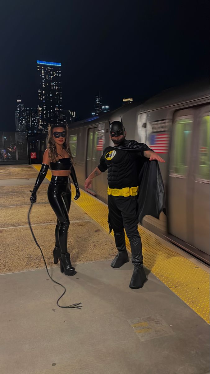 a man and woman dressed up as batman and catwoman on a train platform at night