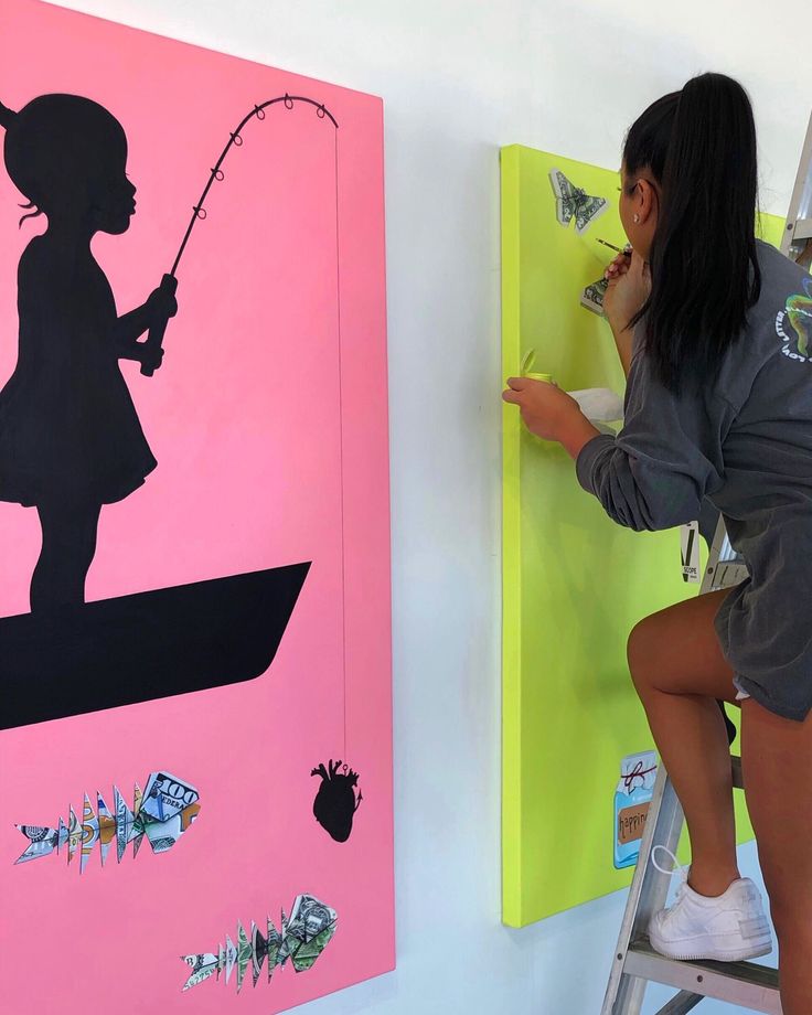 a woman is painting on the wall with pink and yellow paint, while another girl stands next to her holding a fishing rod