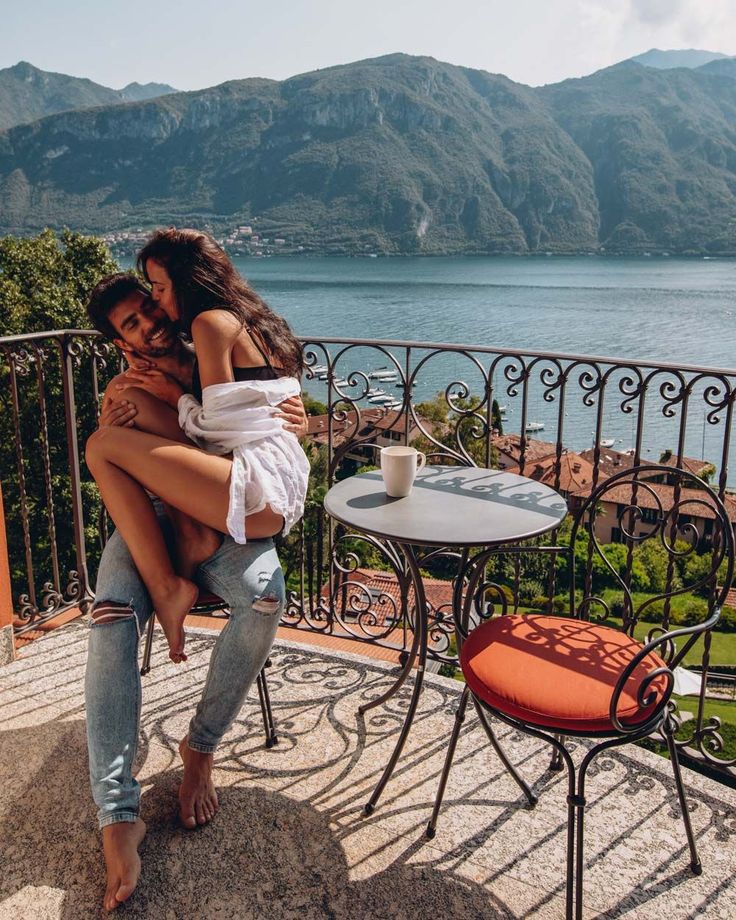 two women hugging each other while sitting at an outdoor table overlooking the water and mountains