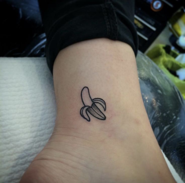 a small banana tattoo on the ankle is shown in black and grey ink, with an outline of two bananas behind it