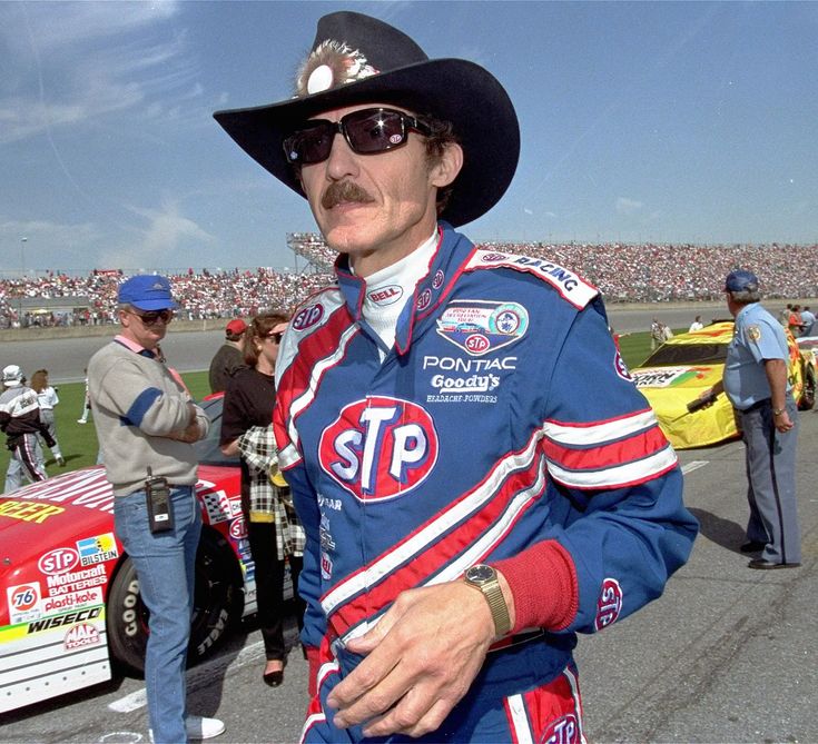 a man in a cowboy hat and sunglasses standing next to a car at a race track