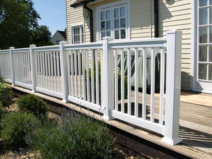 a white picket fence next to a house with flowers in the foreground and bushes on the other side