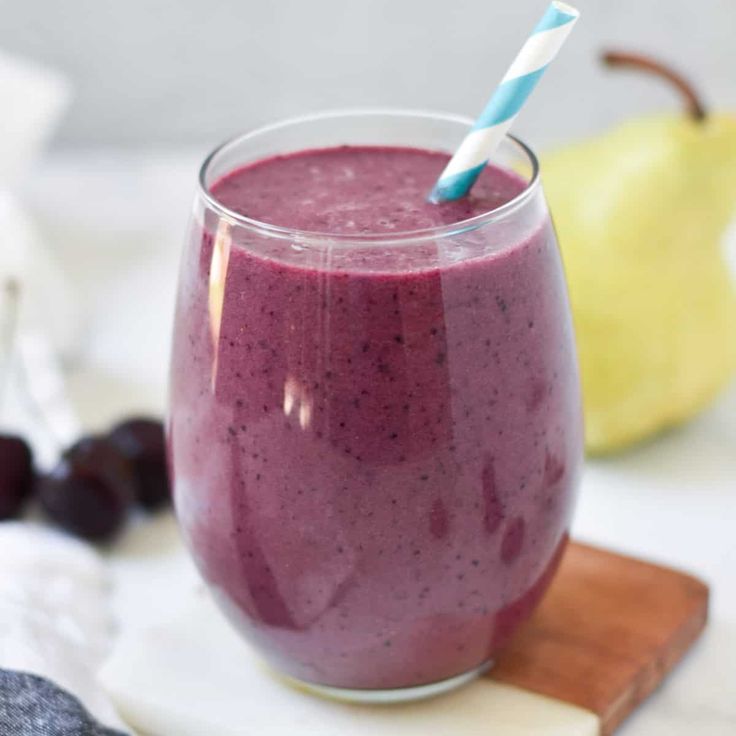 a purple smoothie in a glass with a blue and white striped straw sitting on a cutting board