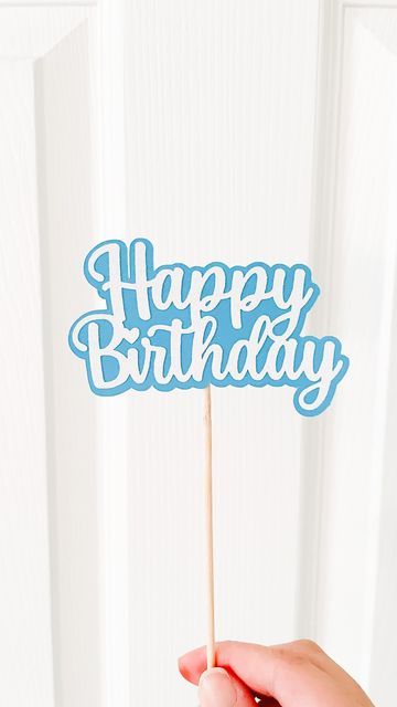 a hand holding a blue happy birthday cake topper in front of a white door