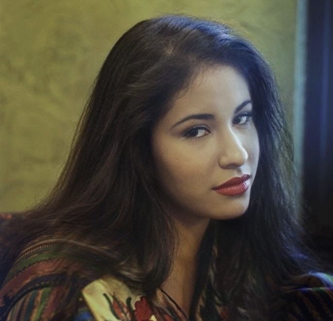 a woman with long hair sitting down and looking off to the side in front of a window