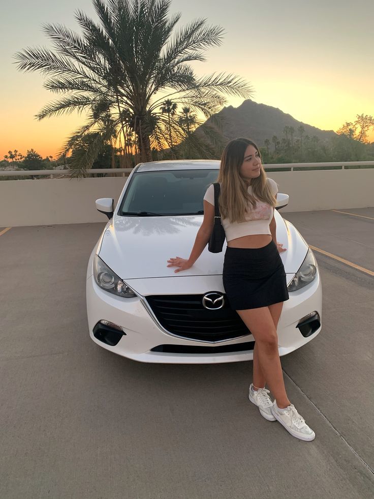 a woman leaning on the hood of a white car in a parking lot at sunset