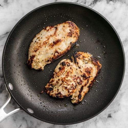 two pieces of chicken cooking in a frying pan on a marble counter top with a spatula