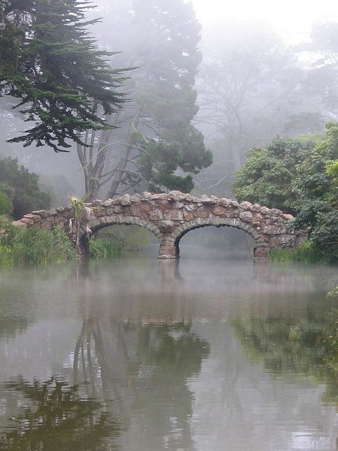 a stone bridge in the middle of a lake surrounded by trees and bushes on a foggy day