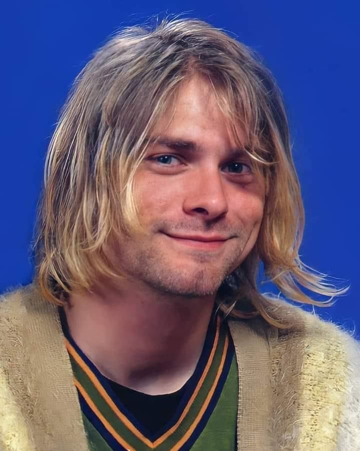 a man with long blonde hair wearing a cardigan sweater and smiling at the camera