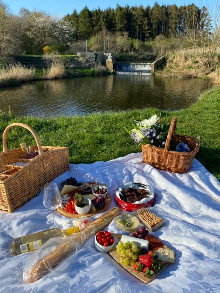 a picnic table with cheese, fruit and bread on the grass near a riverbank