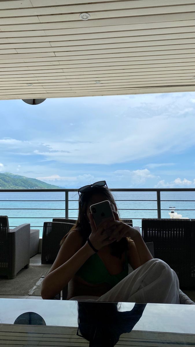 a woman taking a selfie with her cell phone in front of the ocean while sitting on a bench