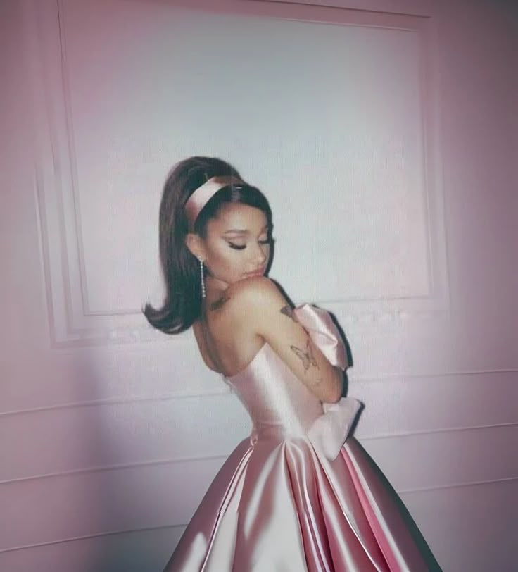 a woman in a pink dress standing next to a wall with her hands on her chest