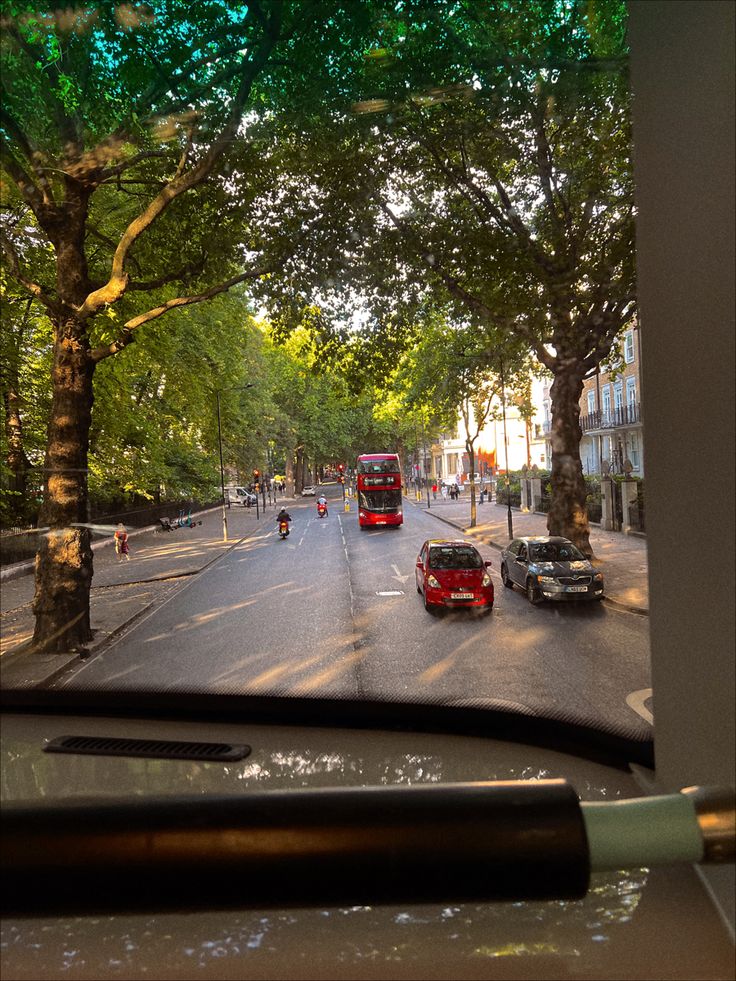 a red double decker bus driving down a street next to tall trees and people walking on the sidewalk