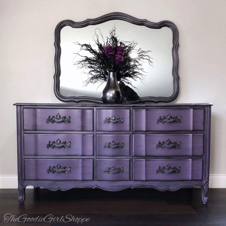 a purple dresser with flowers in a vase on top and a large mirror above it