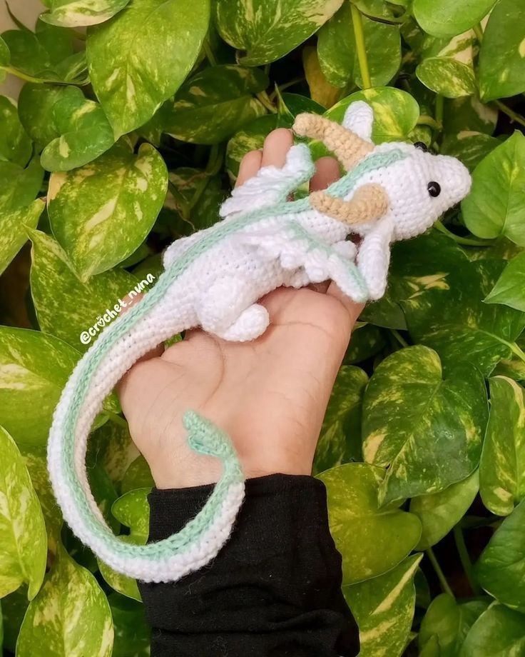 a hand holding a small crocheted toy gecko in front of some green plants