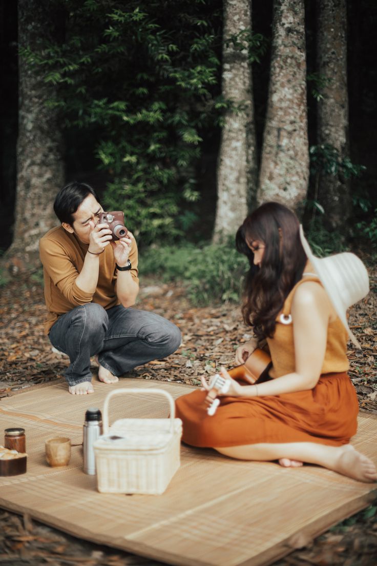 a man and woman sitting on a blanket in the woods, one holding a camera