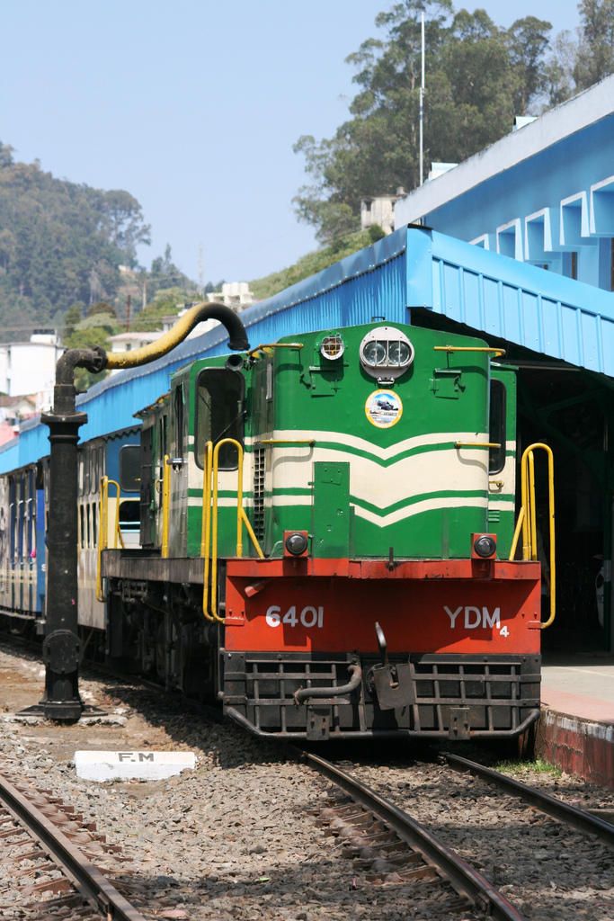 a green and red train traveling past a blue building on railroad tracks with trees in the background