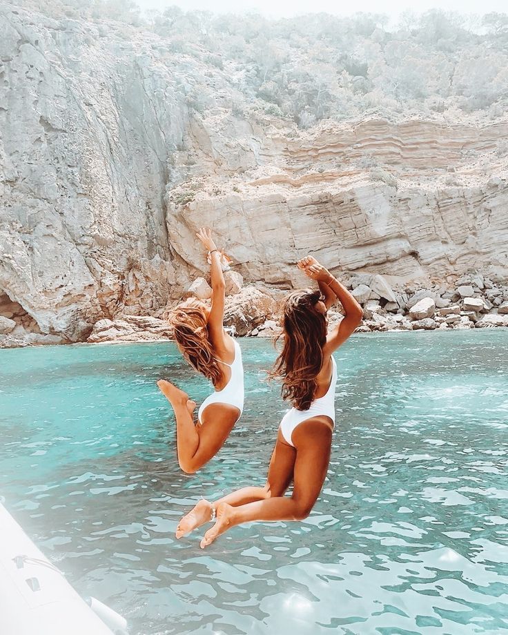 two women in white swimsuits jumping into the water from a boat near a rocky cliff