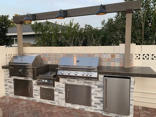 an outdoor kitchen with grills and lights on the outside wall, surrounded by brick pavers