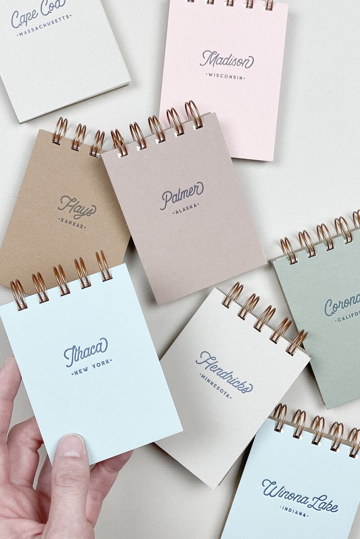 a hand holding a spiral notebook in front of five different colored paper sheets with writing on them