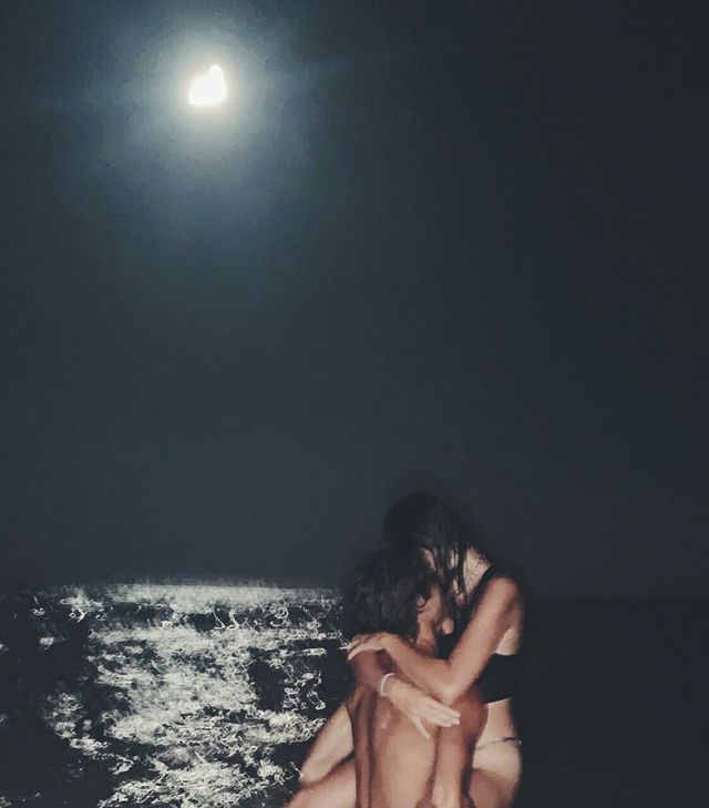 two people are sitting on the beach at night time, one is hugging the other