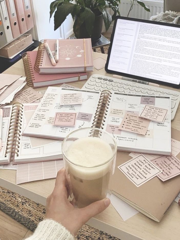 a person holding a drink in front of a laptop on a table with notebooks and papers