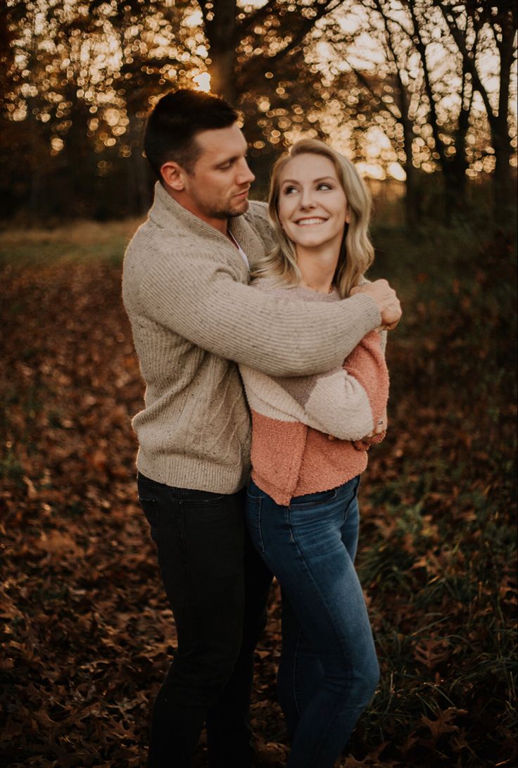 a man and woman hugging each other in the woods at sunset with leaves on the ground