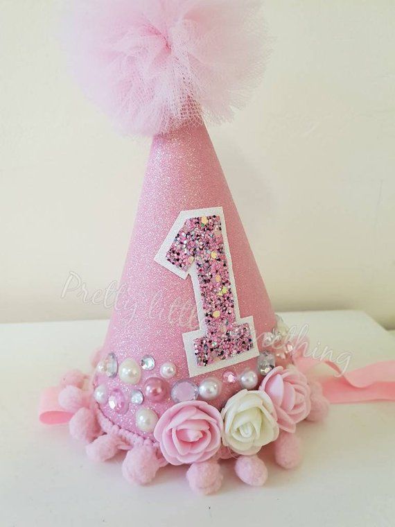 a pink birthday hat with a number one on it and flowers around the top, sitting on a table