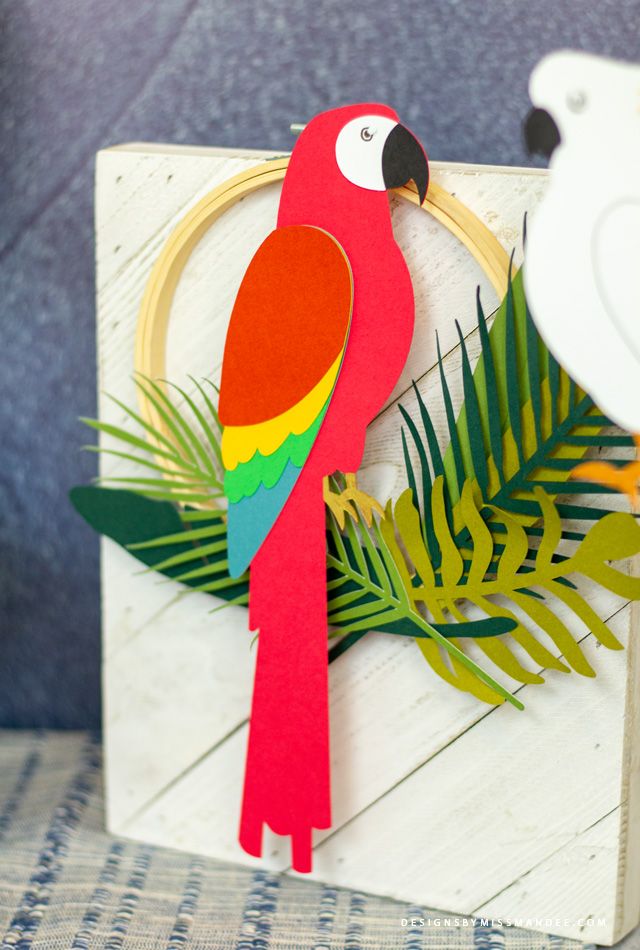 a red parrot sitting on top of a wooden sign next to a bird and palm leaf