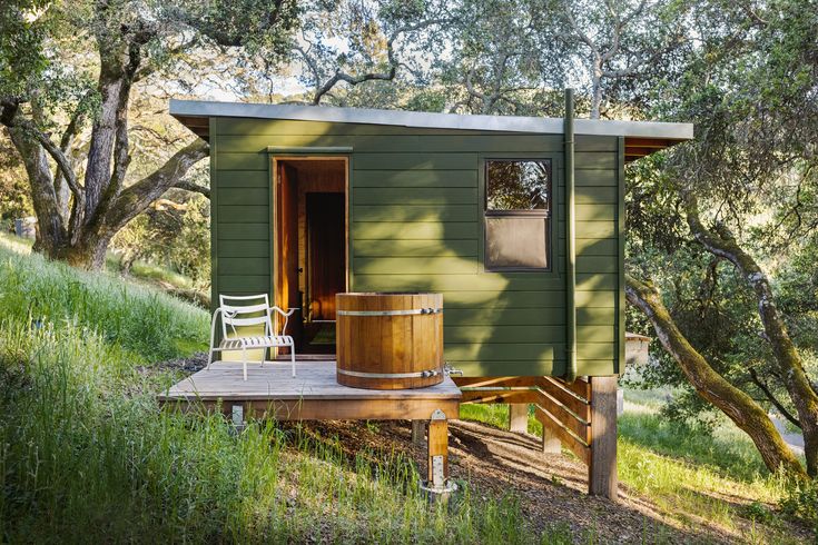 a small green house with a wooden barrel on the porch and trees in the background