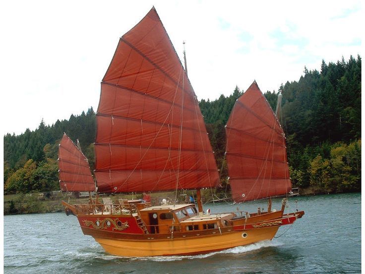 an orange sailboat with red sails on the water in front of trees and hills