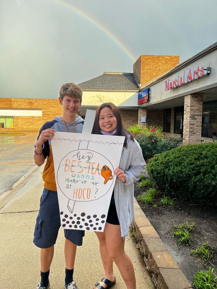 two young people holding up a sign with a rainbow in the sky behind them on a sidewalk